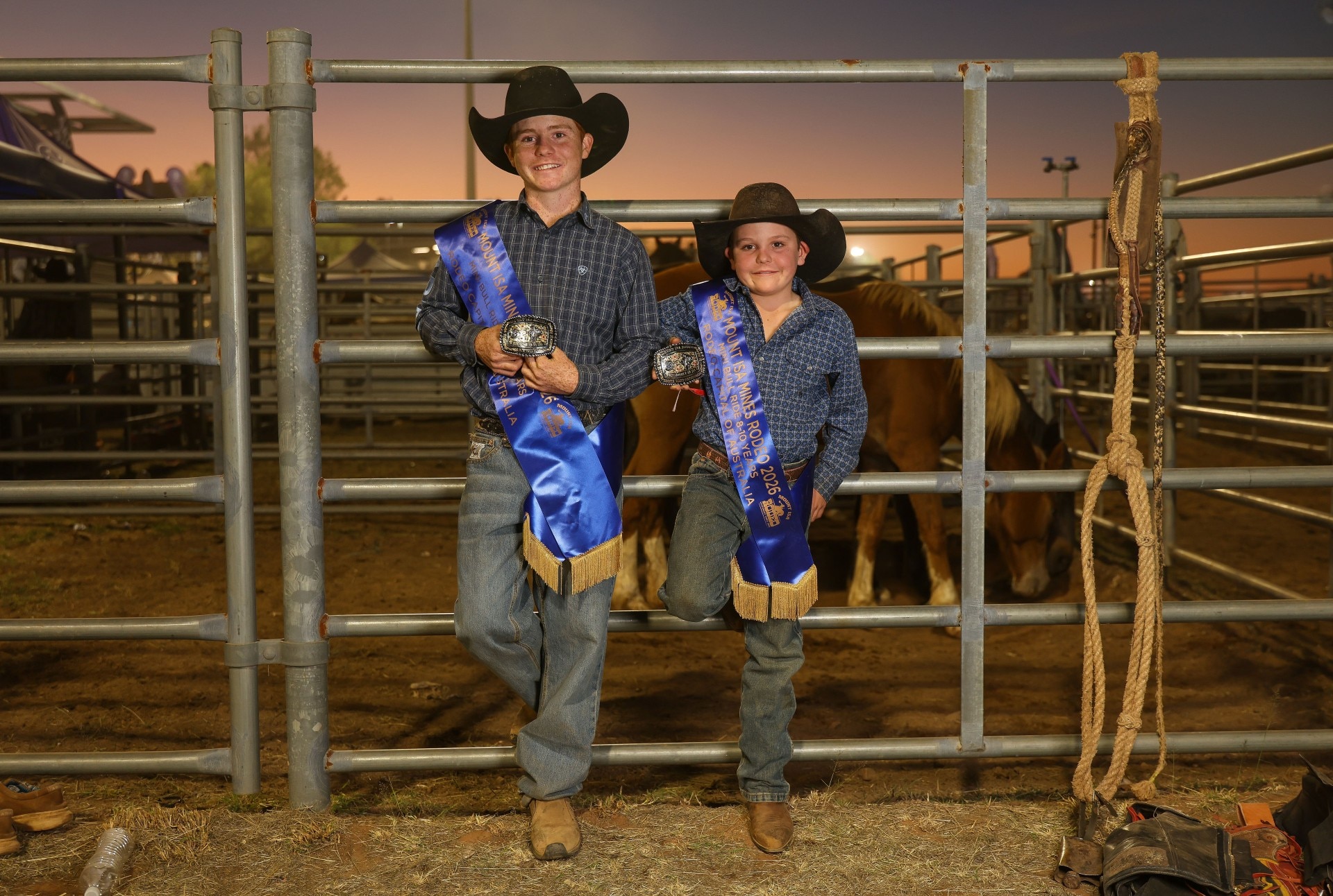 A pair of boys in rodeo gear lean against a fence in a stockyard.