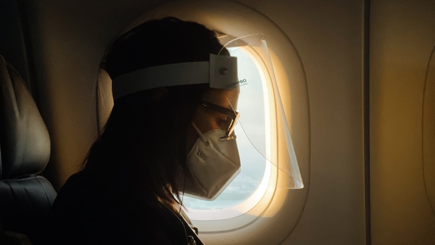 Woman sitting next to airplane window wearing a surgical face mask and face shield 