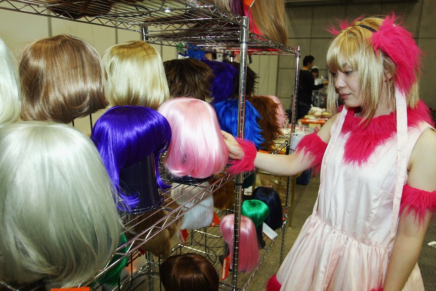 A young woman in a pink and white and a blonde wig handles a bunch of wigs