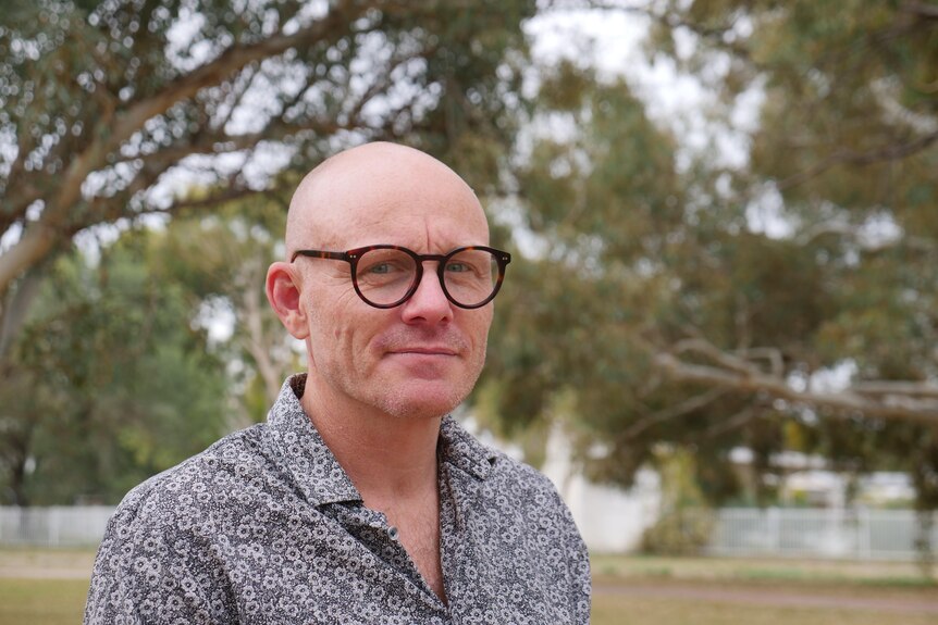 A bald man with glasses smiles softly at the camera. He is standing outside among trees.