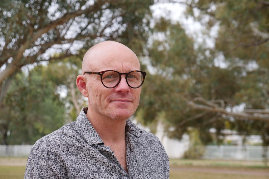A bald man with glasses smiles softly at the camera. He is standing outside among trees.
