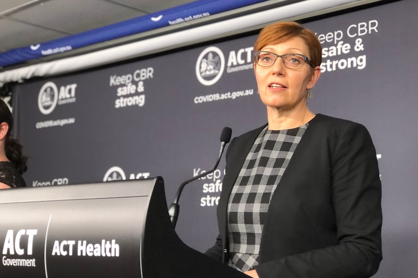 A woman in a check shirt and blazer stands at a podium, delivering a speech.