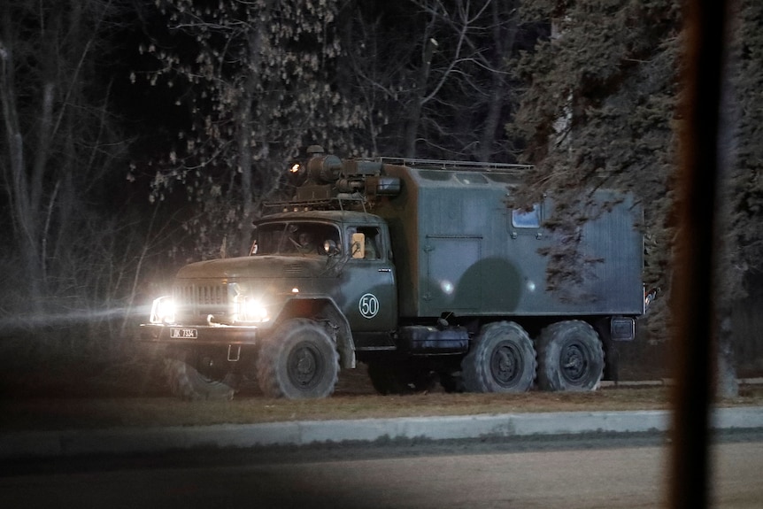 A large dark green truck drives down a dark street with its headlights on