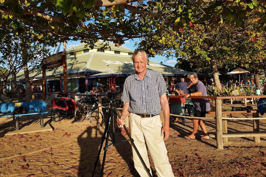 An older gentleman stands in dirt, with a pub behind him and camera on tripod beside him.
