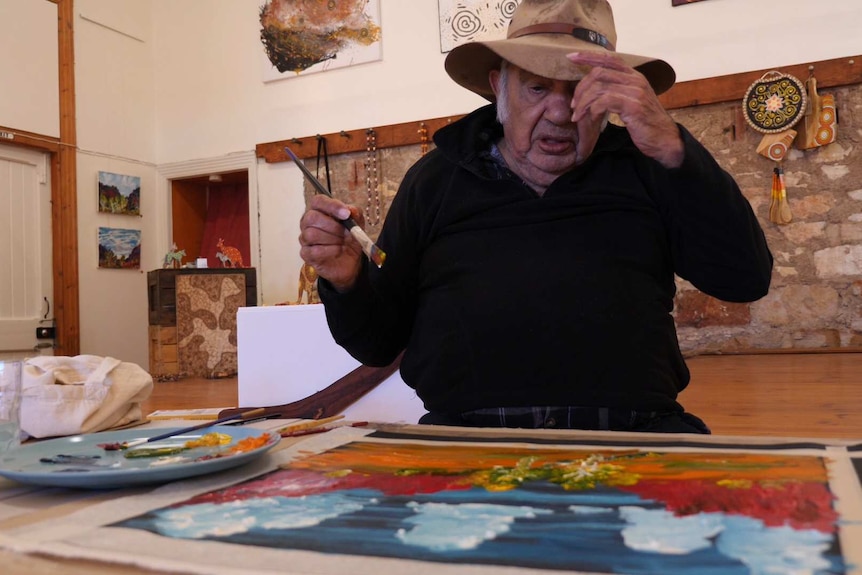 An Aboriginal man in a black jumper sits at a table holding a paint brush.