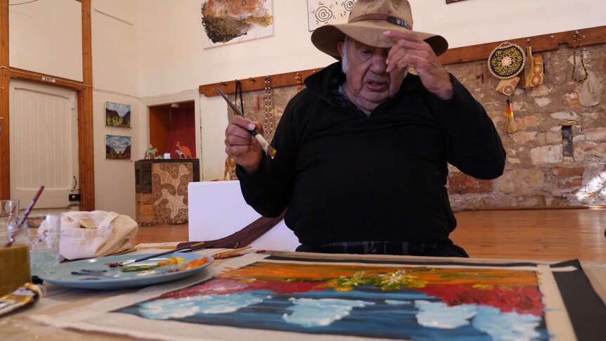 An Aboriginal man in a black jumper sits at a table holding a paint brush.