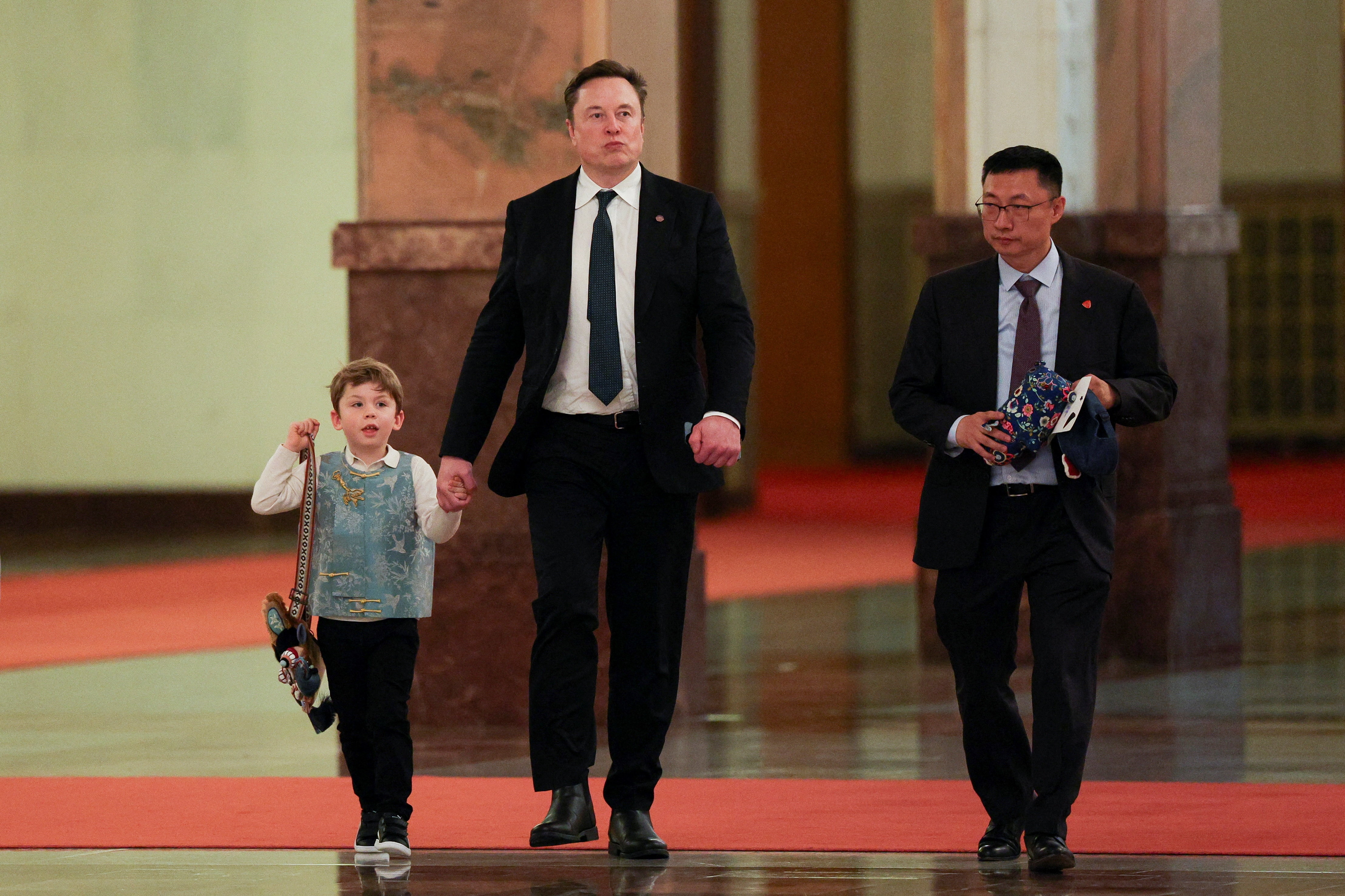 A small child wearing a blue vest walks alongside a man in a suit while holding his hand as a third man walks alongside them