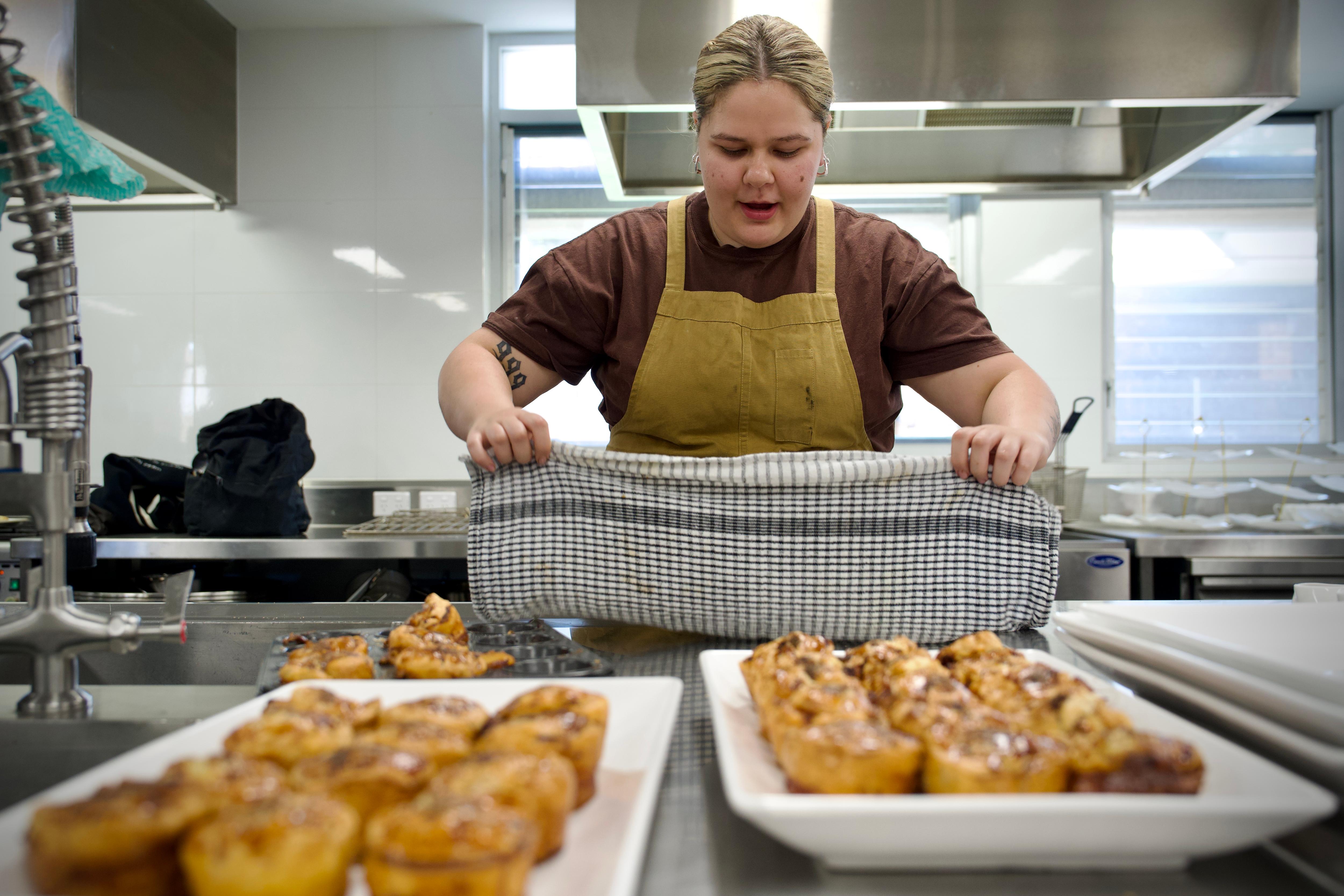 An apprentice Indigenous chef stands in a commercial kitchen holding a tea towel while working on a batch of pies