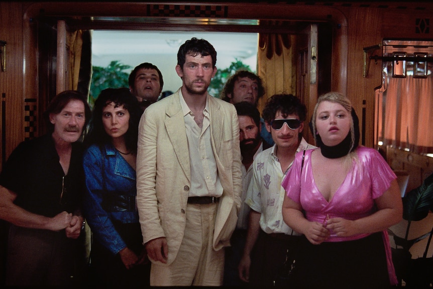 A motley group of characters gather around a man in a linen suit.