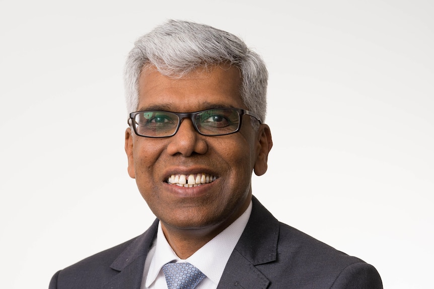 a man in a suit smiles at the camera, standing against a plain white background 