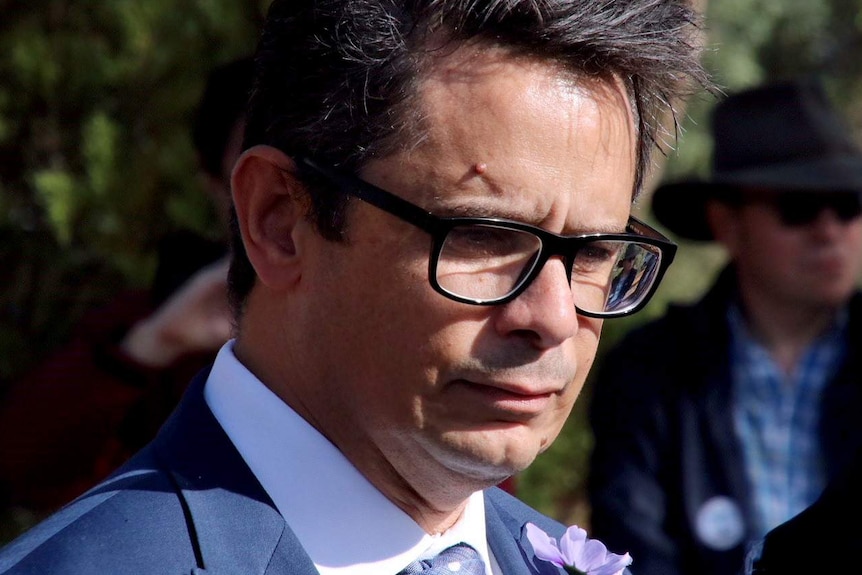 A headshot of a man in a suit and wearing glasses, standing outside in front of trees.