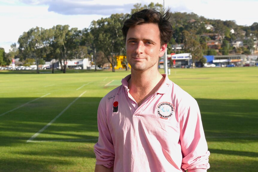 A young, dark-haired man in a cricket uniform stands on the edge of an oval.
