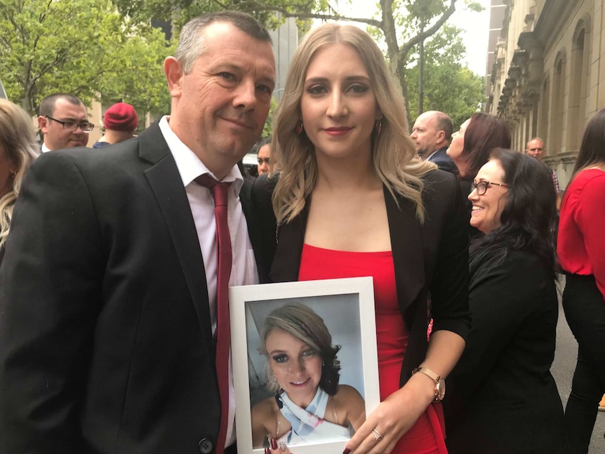 A man and woman dressed in red stand outside a court building with a photo of a young woman.