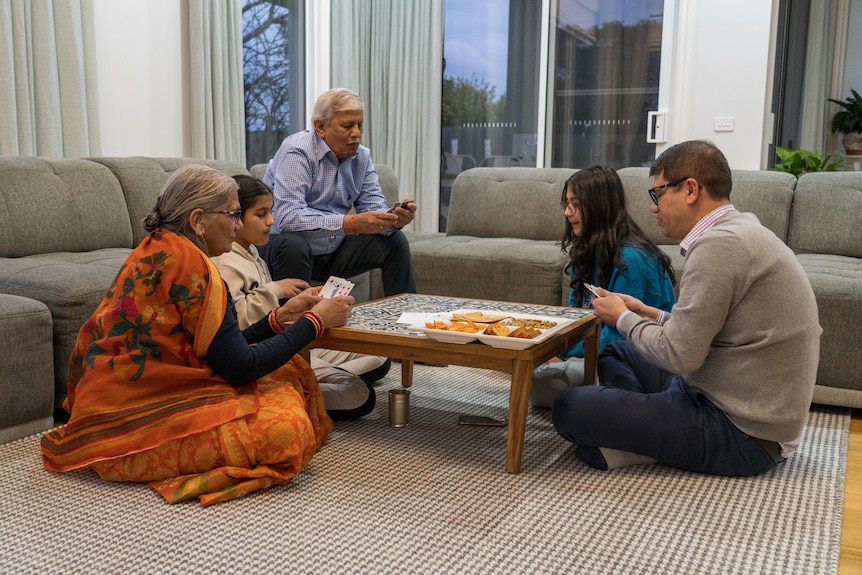 A father, his two children and his parents sit around a coffee table playing cards. There is a plate of snacks on the table. 