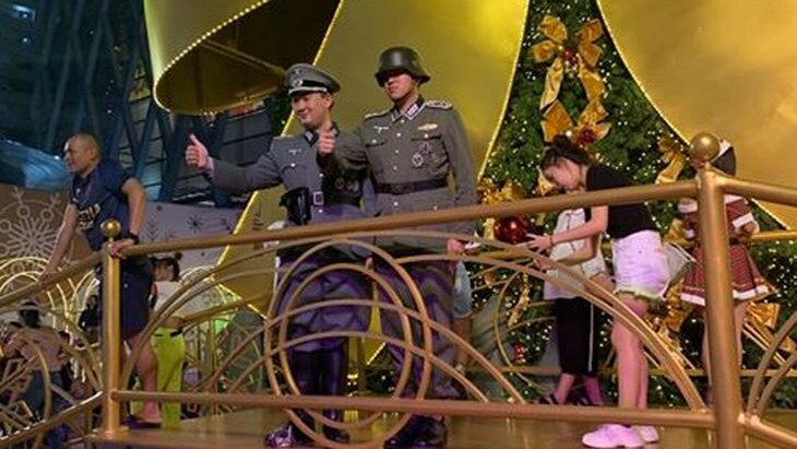 Two men stand on a large Christmas-themed stage dressed in Nazi military uniforms and give the thumbs-up to onlookers.