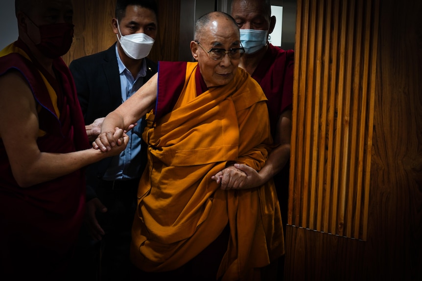 The Dalai Lama in his trademark orange and maroon robes is supported by several minders propping him up
