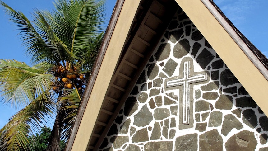 The outside top of a stone church with a cross in the stonework and a palm tree behind.