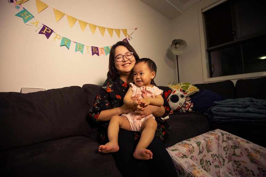 A mother gives her baby daughter a big hug on the couch at home. Mother mother and daughter smile with glee.