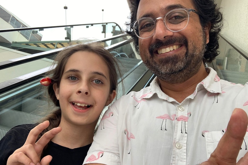 A man wearing glasses and a beard stands on an escalator with a young girl. 