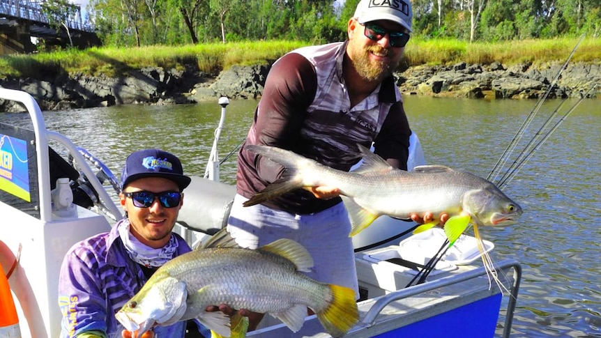 Two men wearing caps and sunglasses hold a fish each over the edge of a charter fishing boat in a river