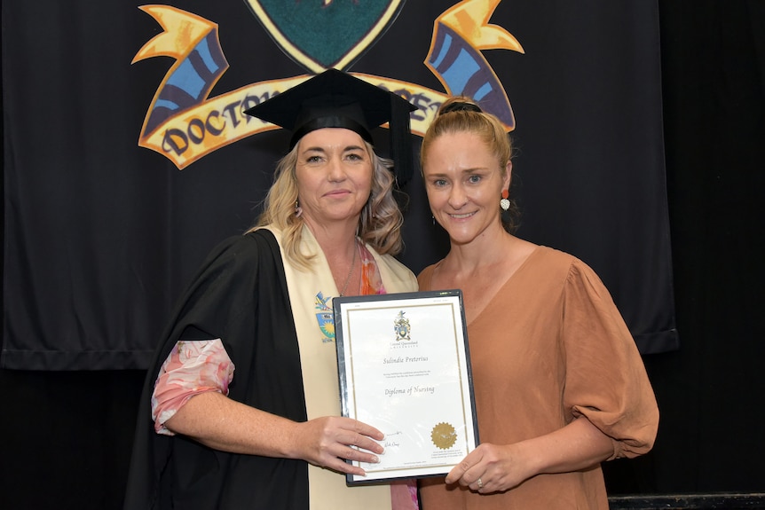 A woman in a graduation gown smiling and holding a diploma, next to another woman smiling in front of a university banner.