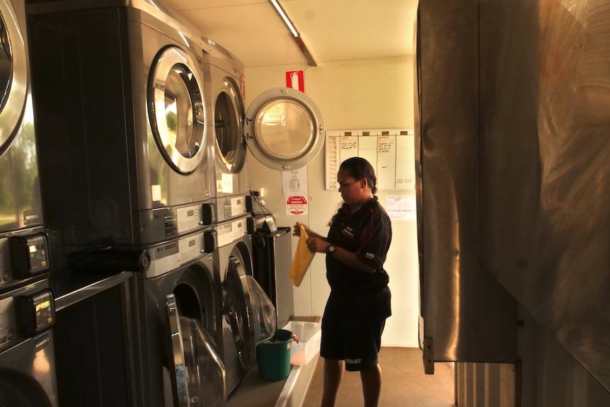 A woman holds a yellow teatowel in front of a row of industrial sized washing machines. 