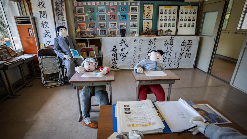 Two dolls dressed in children's clothes are propped at desks while a doll in a teacher costume sits at a front desk.