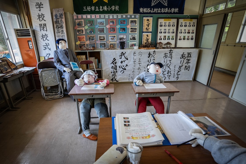 Two dolls dressed in children's clothes are propped at desks while a doll in a teacher costume sits at a front desk.