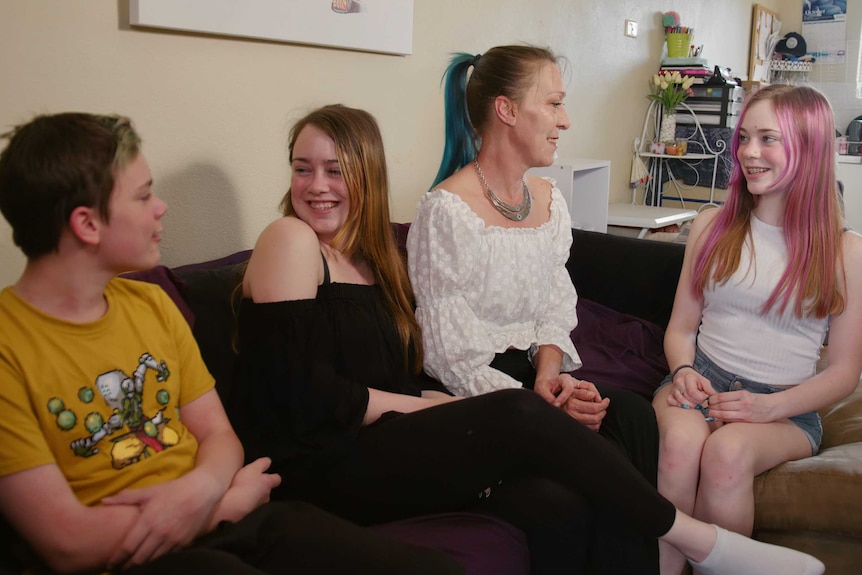 a mother and her three teenagers sit on a couch, chatting happily.
