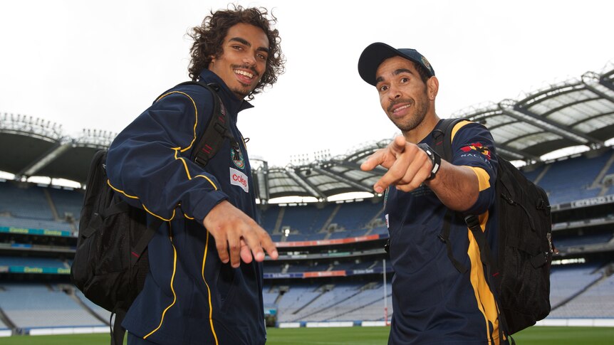 Tony Armstrong and Eddie Betts, wearing Australian training tracksuits, smile and point at the camera