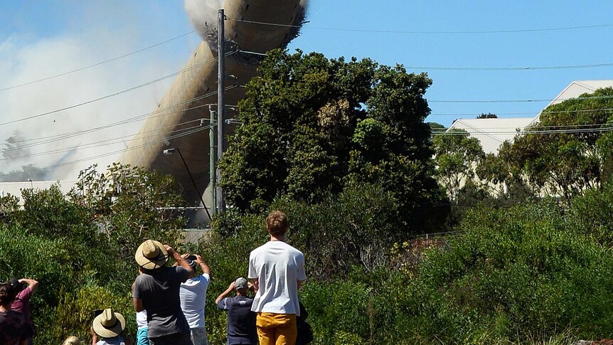 Wide shot of people watching on as a high smoke stack tower falls towards the ground.