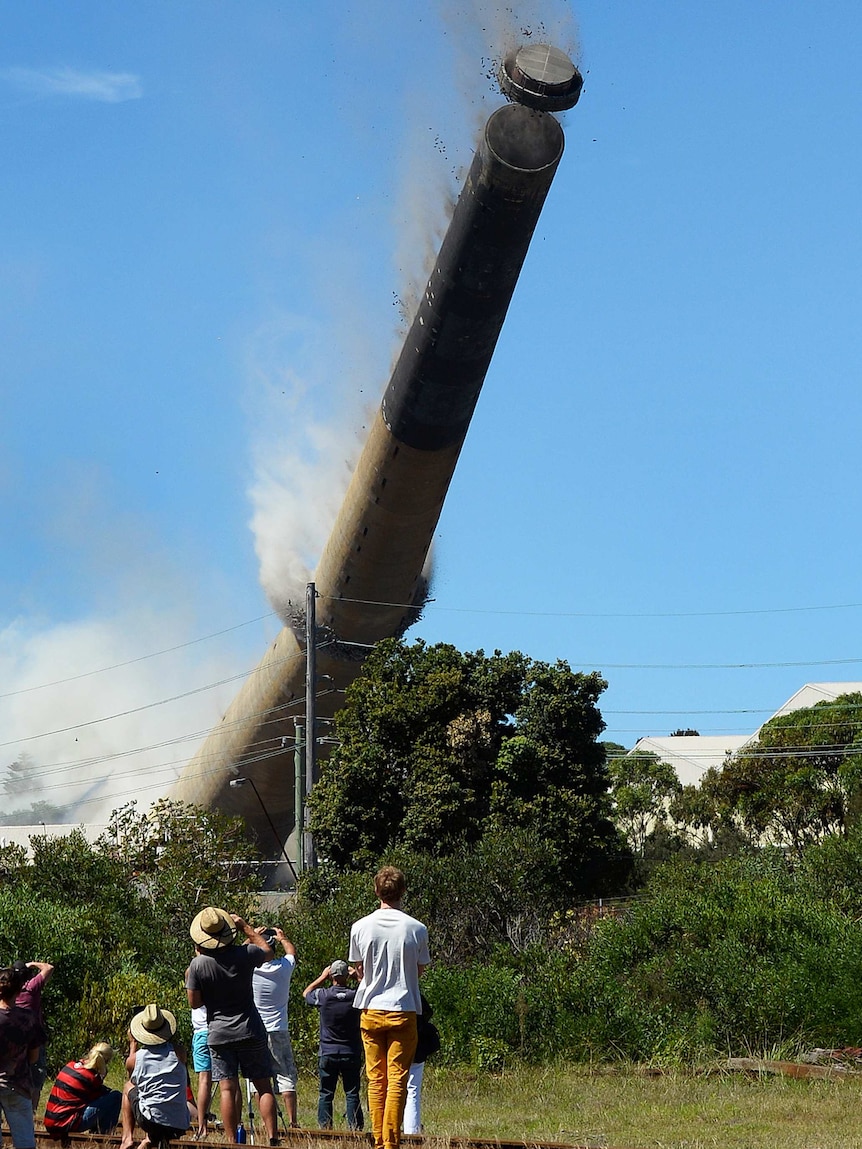 Wide shot of people watching on as a high smoke stack tower falls towards the ground.