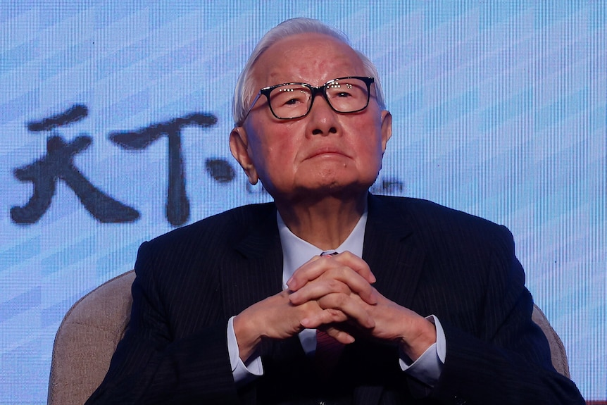 Man wearing glasses in a suit looks serious with hands clasped in front of his chest.