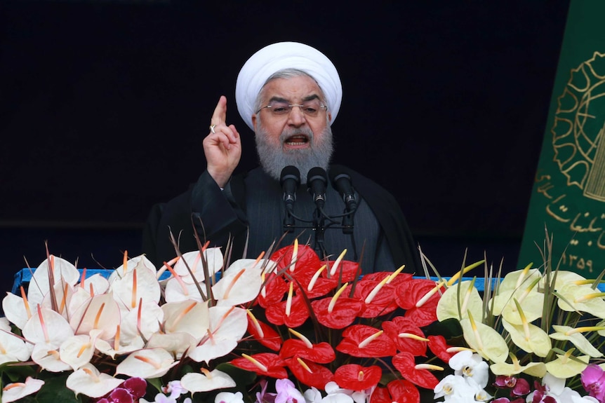 President Rouhani points his right index finger speaking on platform adorned with red, pink, and yellow Anthurium flowers.
