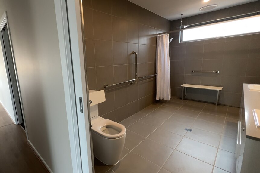 Large bathroom with grey floor and wall tiles, grab rails and a white bench seat.