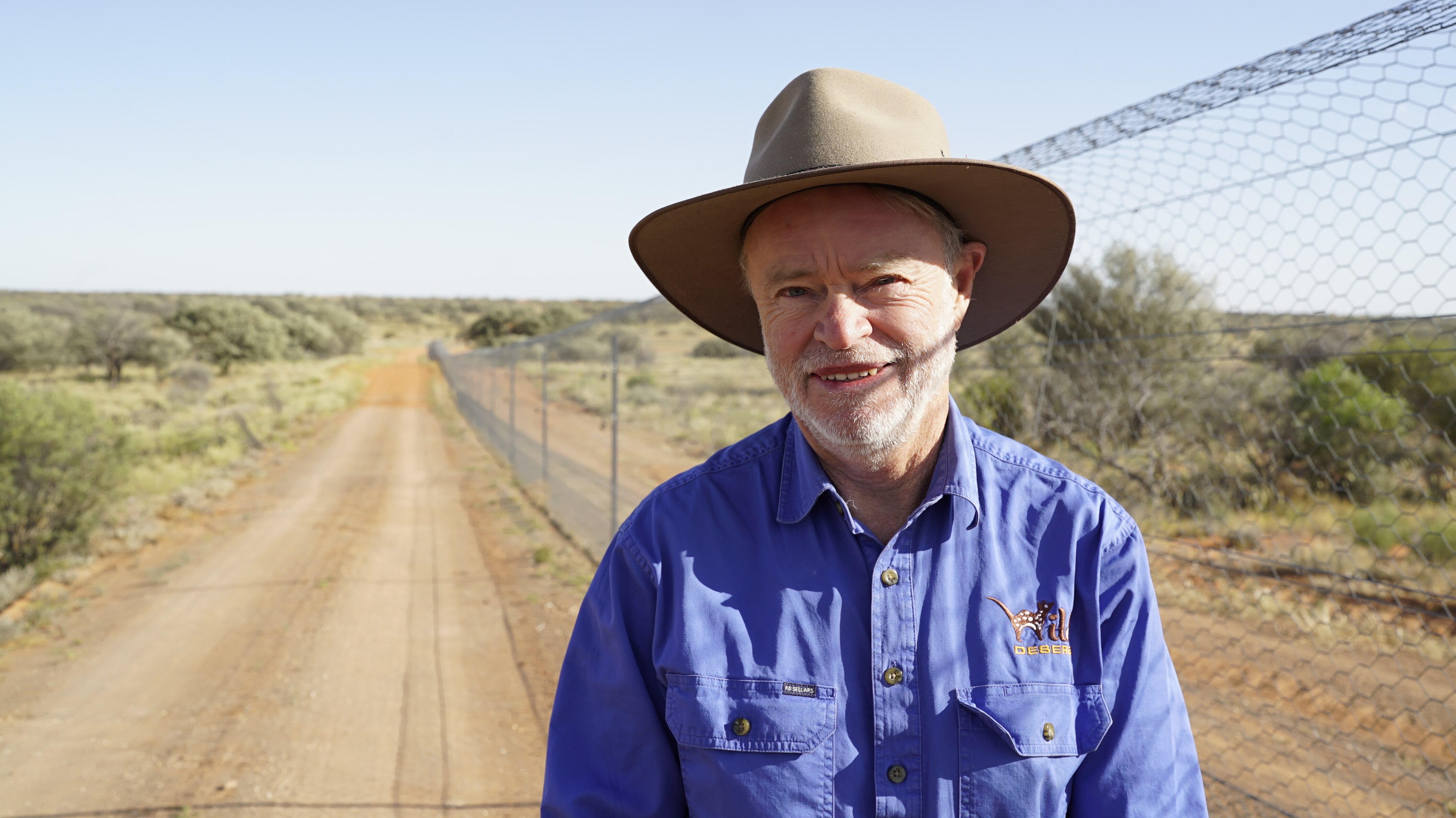 Man in a blue shirt and hat standing next to a fence running through the desert.