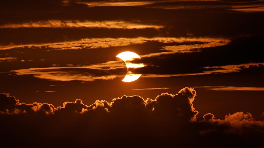 A red-and-orange-hued photo of the sun partially hidden behind clouds.
