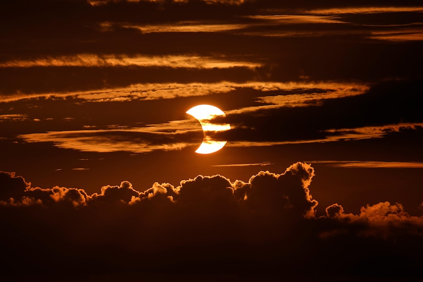 A red-and-orange-hued photo of the sun partially hidden behind clouds.