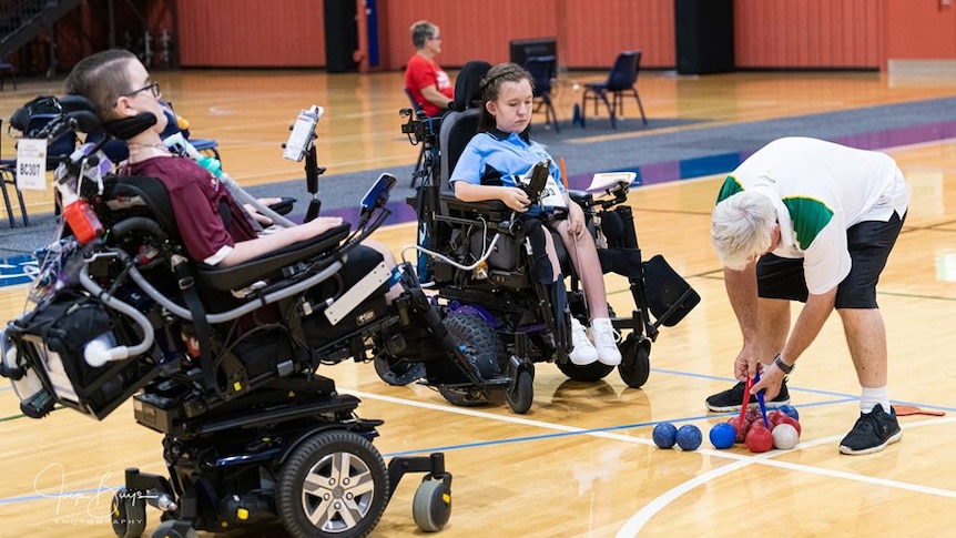 Two young people with disabilities play a ball game from their wheelchairs. A referee measures the distance between the balls.