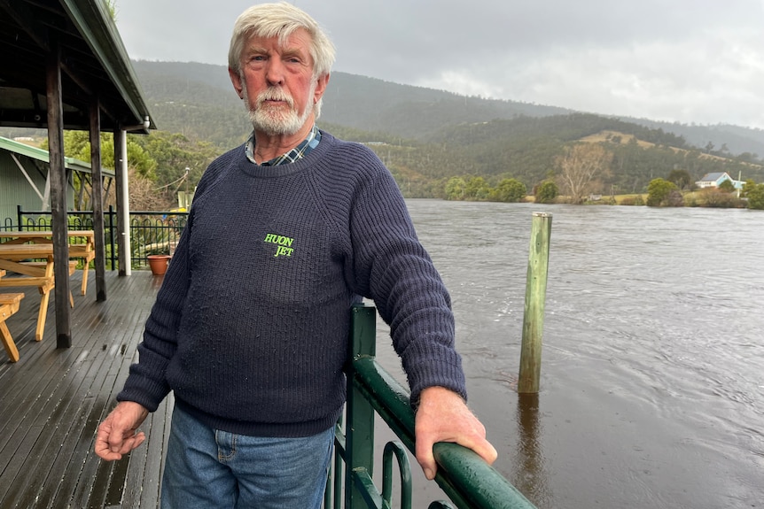 Man with white hair and a beard stands on the deck of his business with the flooded Huon River behind him