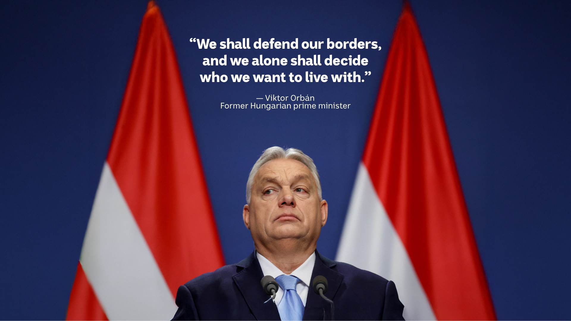 A white man with grey hair wears a black suit and light blue tie, with two large Hungarian flags behind.