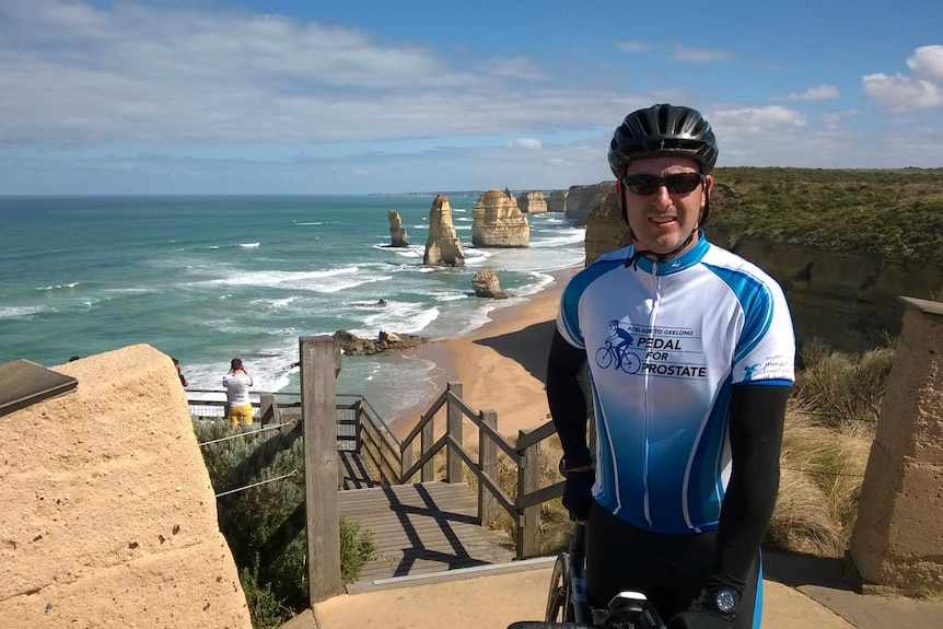 A man in cycling clothing and a helmet stands with the Twelve Apostles behind him