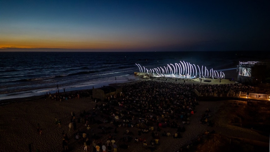 A crowd of people gather in front of a lit-up stage on a darkened beach after sunset.