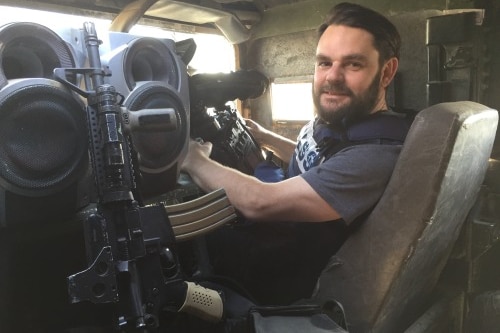 Aaron sits inside a military vehicle, next to a gun, as he turns back to face the camera.