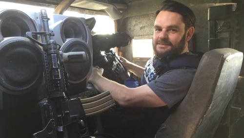 Aaron sits inside a military vehicle, next to a gun, as he turns back to face the camera.