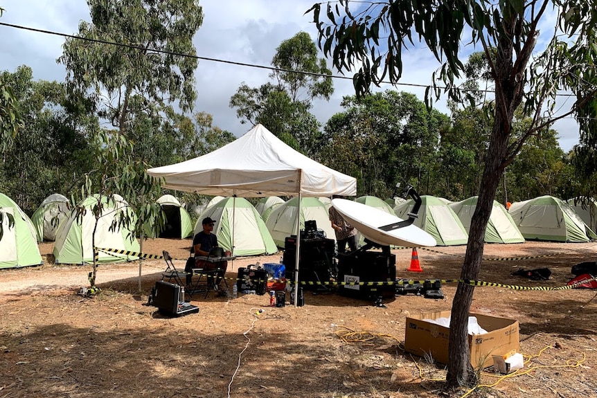 Satellite dish and equipment under small marquee set up in front of row of tents in bush.