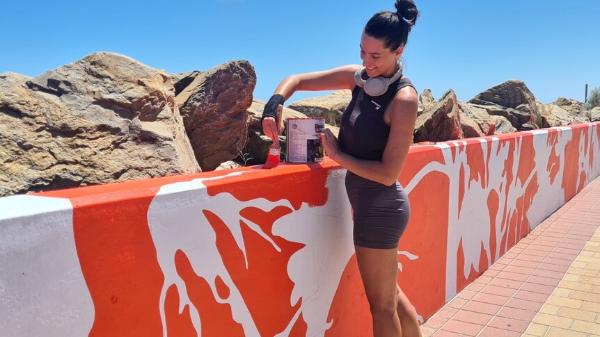 woman in black gym gear with grey headphones around her neck paints a seawall in orange and white with rocks in background