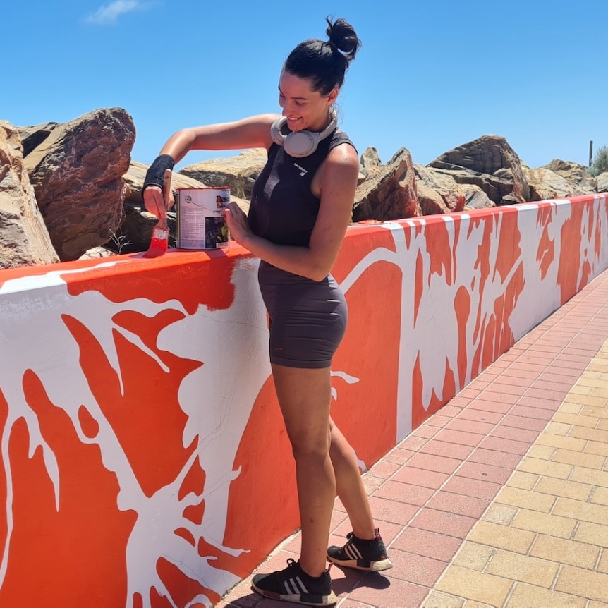 woman in black gym gear with grey headphones around her neck paints a seawall in orange and white with rocks in background