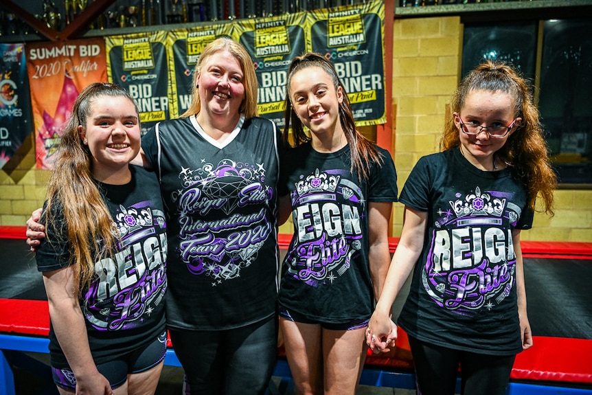 Four women look and smile at the camera in a cheerleading gym.