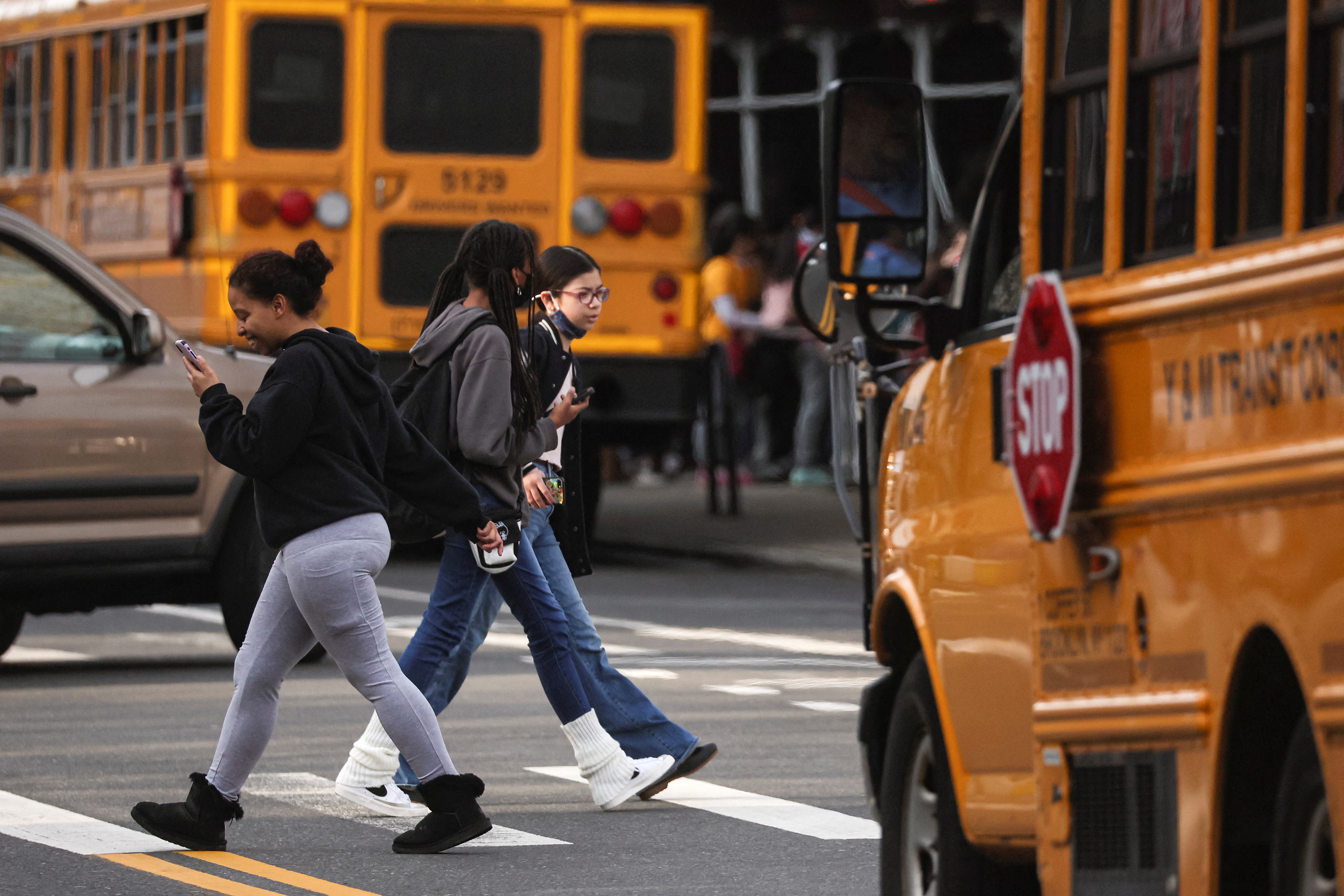 Children walk across a pedestrian crossing past multiple yellow school buses. 